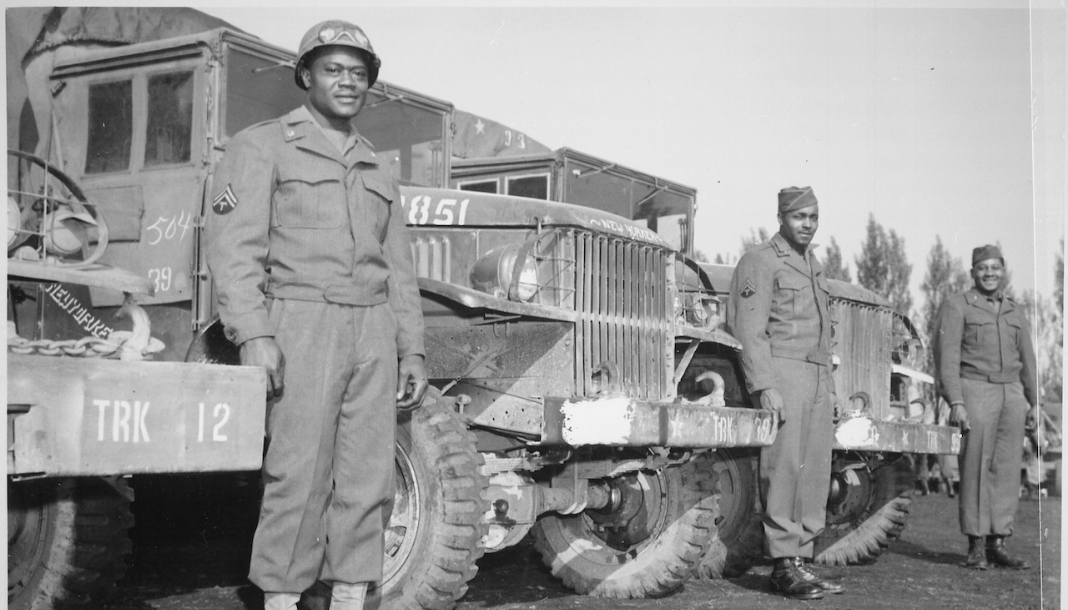 Black and white historical photo of World War Two African American soldiers standing next to military vehicles. 