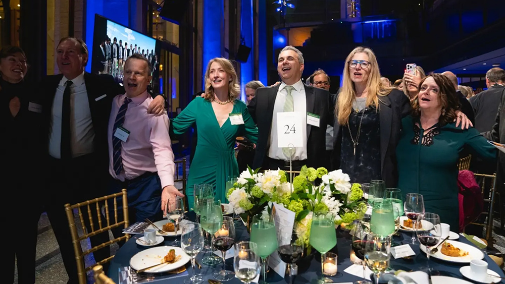 From left are Annie Sundberg ’90, Senior Vice President for University Advancement Bob Lasher ’88, Chris Kelley ’88, Julia Lazarus ’90, Jeff Lazarus ’87, Ricki Stern ’87, and Rachel Dratch ’88 standing singing the alma mater at a table