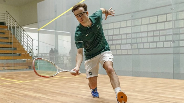 A Dartmouth squash player bends down to swing at a ball coming in low against the ground