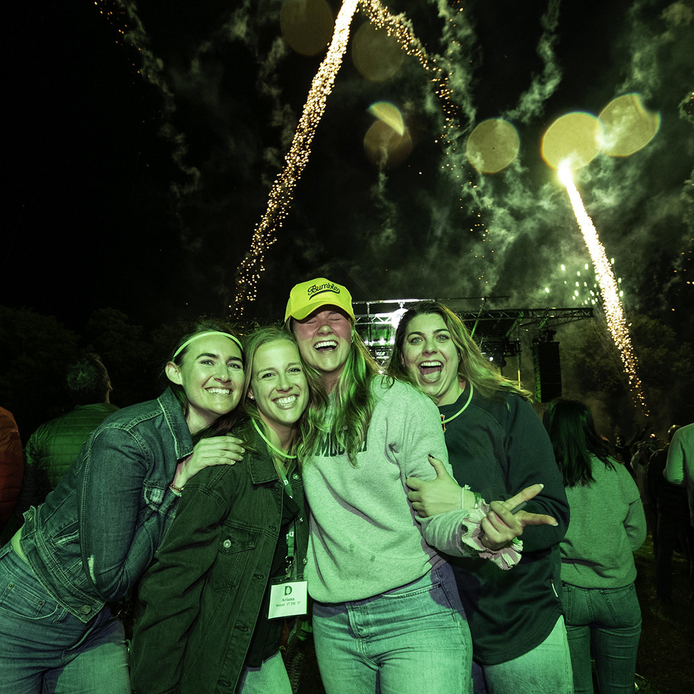 Four students smile at the camera as fireworks go off during a concert on the Green during Reunions