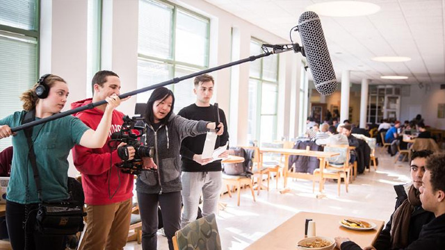 Four students working on a video project holding a boom mic, camera, and other equipment while shooting a video