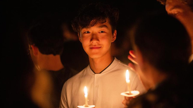 A student, face aglow from candlelight, holding a candle at a vigil on campus