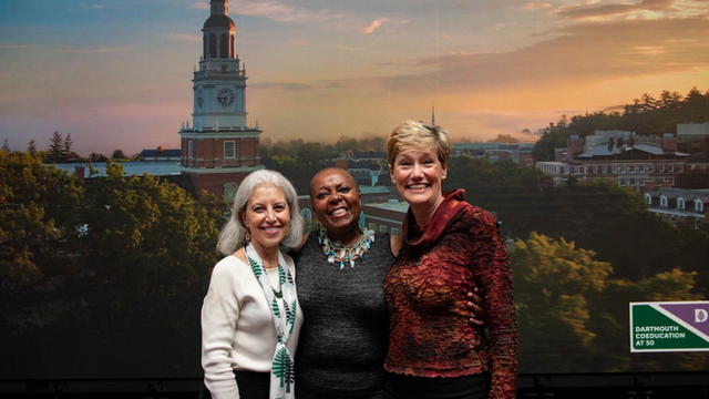 Three women standing arms around each other in front of a backdrop image of Baker Tower and the Coeducation at 50 celebration logo.