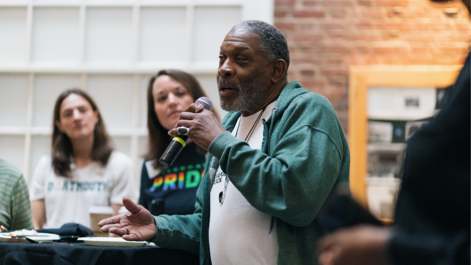 A person in a green hoodie speaking into a microphone at the DGALA Breakfast in 2023.