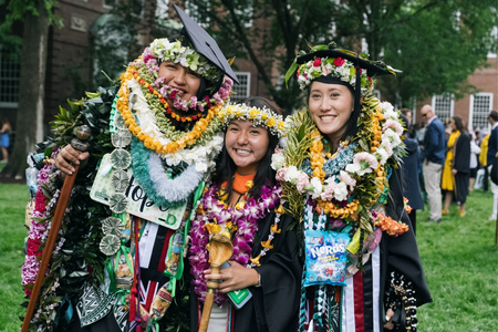 Three graduates smiling and wearing many flower leis and headbands with their regalia. 