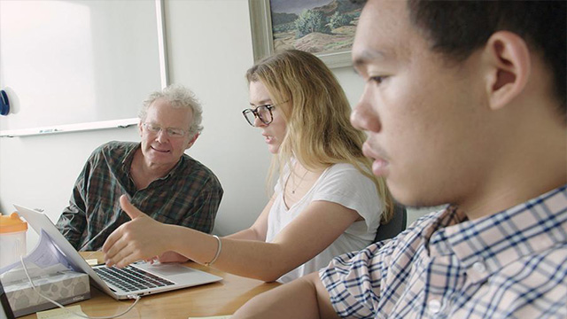 A professor sitting next to two students, assisting one student who is showing him something on a laptop.