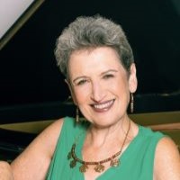 Headshot of Professor Sally Pinkas wearing a green sleeveless blouse, a brown beaded necklace, smiling towards the camera while leaning on a piano behind her.