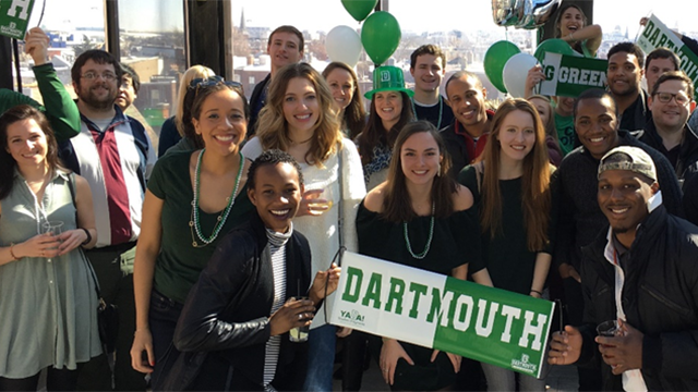 A group of young alumni posing for a photo at an event with green and white balloons and pull out signs that say Dartmouth