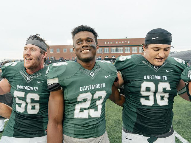 Three football student-athletes standing arm-in-arm on the football field during a Homecoming game.