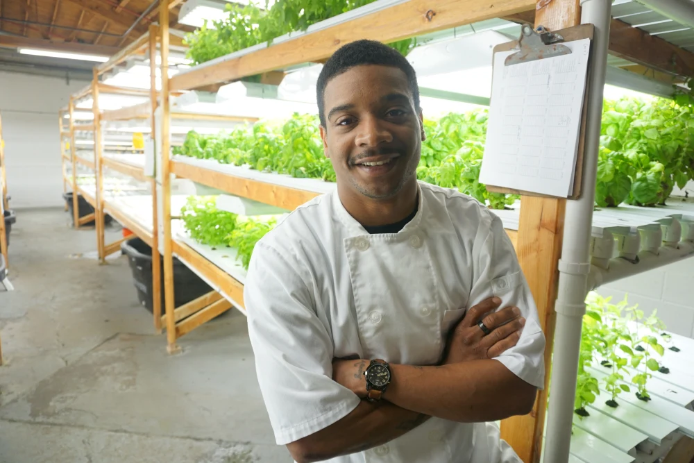 Darius Jones standing in a white t shirt with arms folded and smiling as he stands in front of microgreens plants.