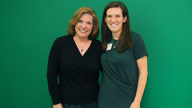 A photo of Katie Paxton, on the right, posing for a photo in front of a green background. 