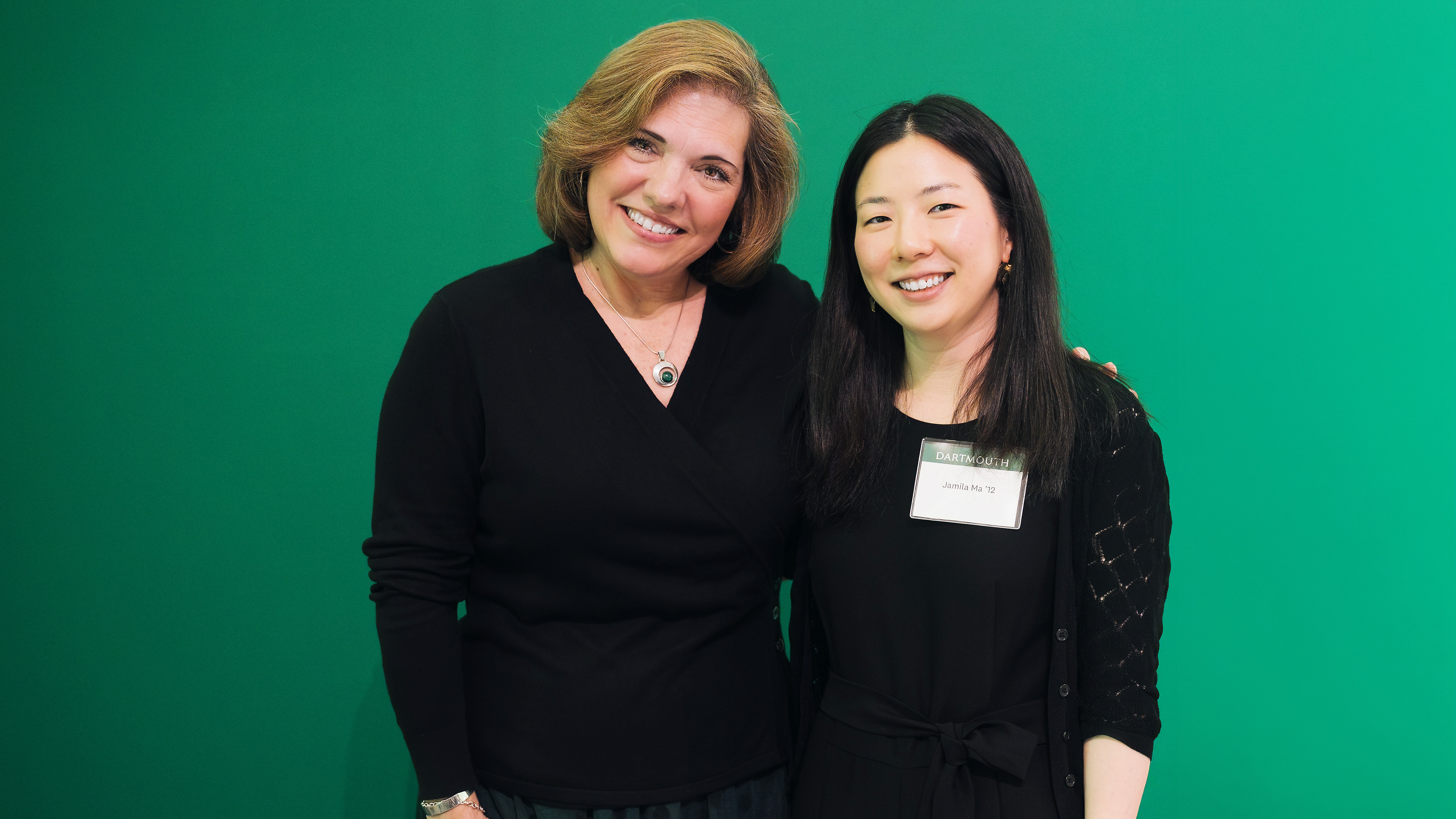 A photo of Jamila Ma, on the right, posing for a photo in front of a green background.