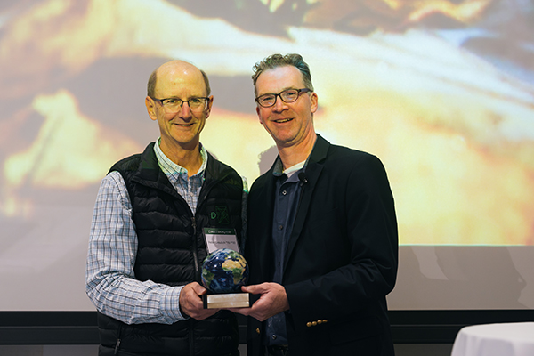 A photo of Professor Jeremy DeSilva posing for a photo, on the right, holding his award 