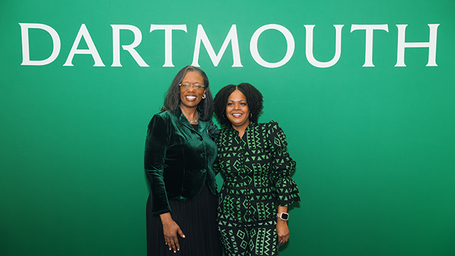 A photo of Crystal Crawford, on the left, posing for a photo in front of a green background with the word Dartmouth on it. 