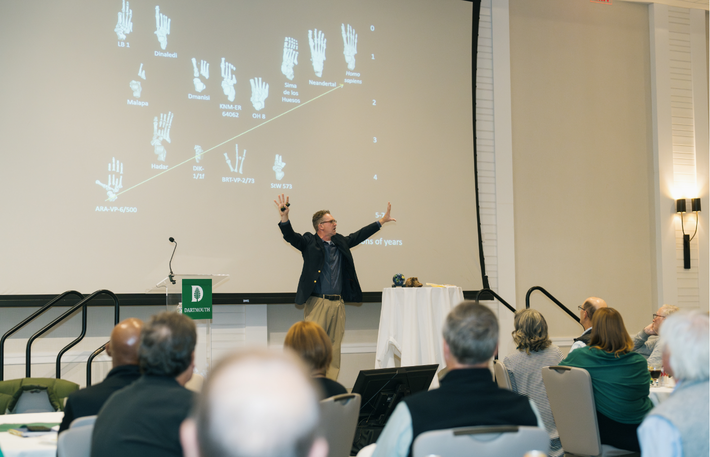 Professor of Anthropology Jerry DeSilva giving a presentation, his arms outstretched as he speaks.