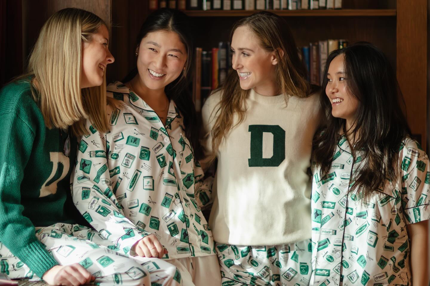 Four women in Dartmouth branded pajamas and Dartmouth sweaters posing in the library. 