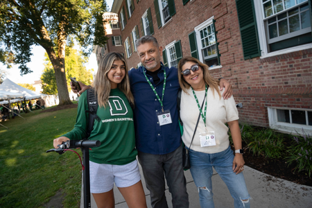 A family wearing their weekend lanyards posing next to a residence hall. The student is wearing a Dartmouth sweatshirt and holding a scooter. 