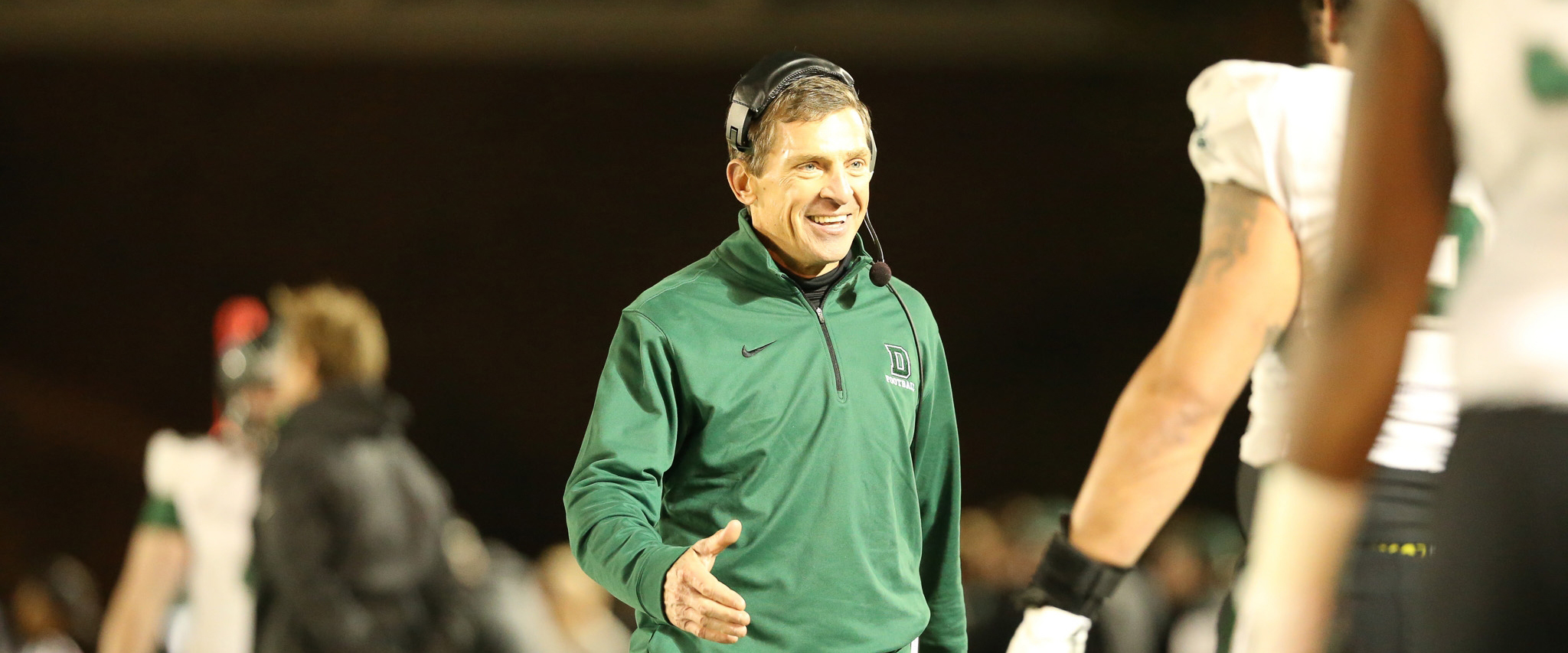 A photo of the late Buddy Teevans on the football field walking toward a student-athlete, smiling, with his hand extended for a hand shake.