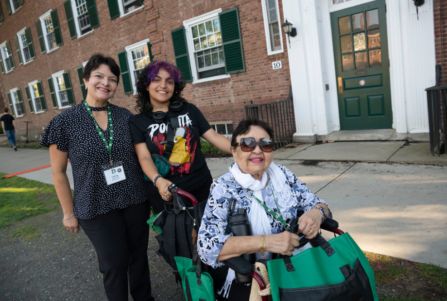 An intergenerational family posing in front of a residence hall. The student has purple streaked hair and headphones, posing with two family members, including a senior seated in a wheelchair