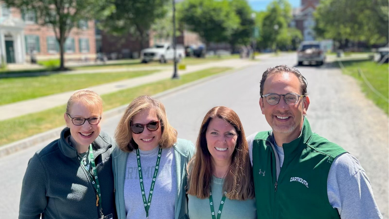 Four alumni council members in Dartmouth gear posing together on Tuck Drive. 