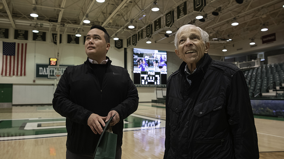 A photo of Dartmouth athletic director Mike Harrity standing next to Stephen Lewinstein, as he gazes into the stands on the basketball court inside the newly renamed Lewinstein Athletic Center