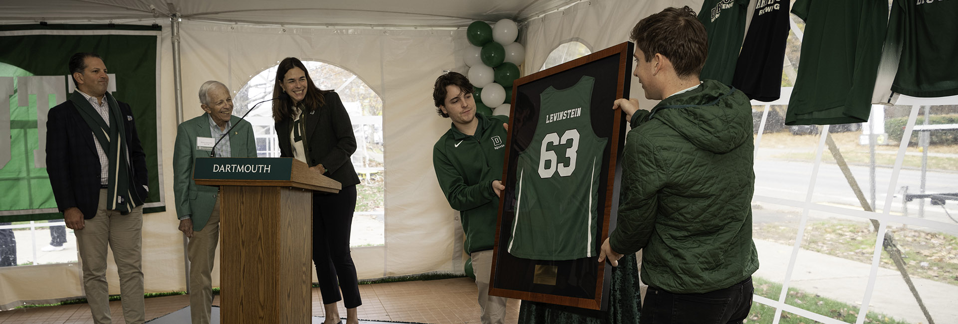 Two student athletes presenting Stephen Lewinstein with a named basketball jersey. Stephen is at the podium with his son and President Beilock