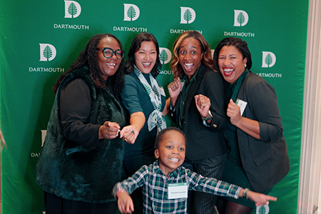 A group of alumni, staff, and family take a photo in front of a Dartmouth banner