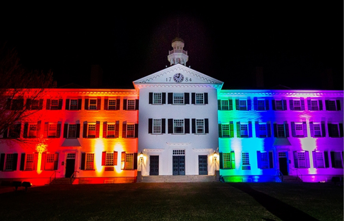 A photo of Dartmouth Hall at night lit up with rainbow colored lights.