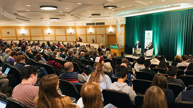 A group of people seated in an auditorium watching two speakers having a discussion on stage.