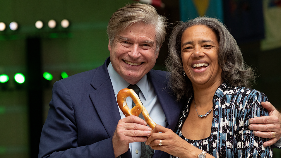 A photo of Don Pease (left) with his arm around Cheryl Bascomb (right). The pair are posing together, smiling as they hold a soft pretzel at an event in Philadelphia.