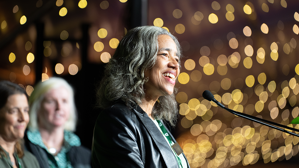 A photo of Cheryl Bascomb smiling as she speaks at a podium during an event.