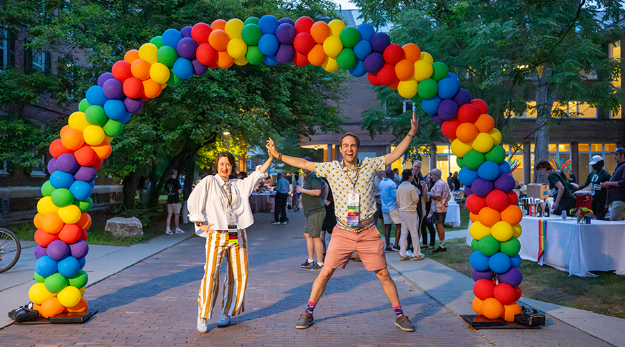 Two people standing under a rainbow balloon arch.