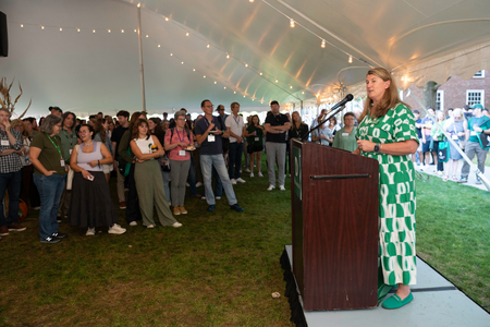A presenter in a green dress speaking at a lecturn under a tent.