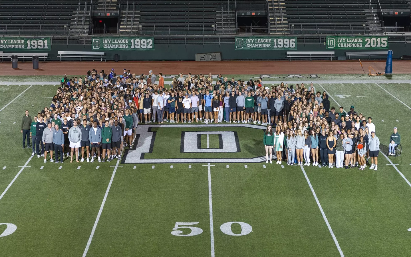 An aerial image of student-athletes gathered around the "D" at the 50-yard line of the football field.