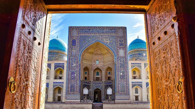 view of an ornate Uzbek madrasah with blue domes through a carved doorway